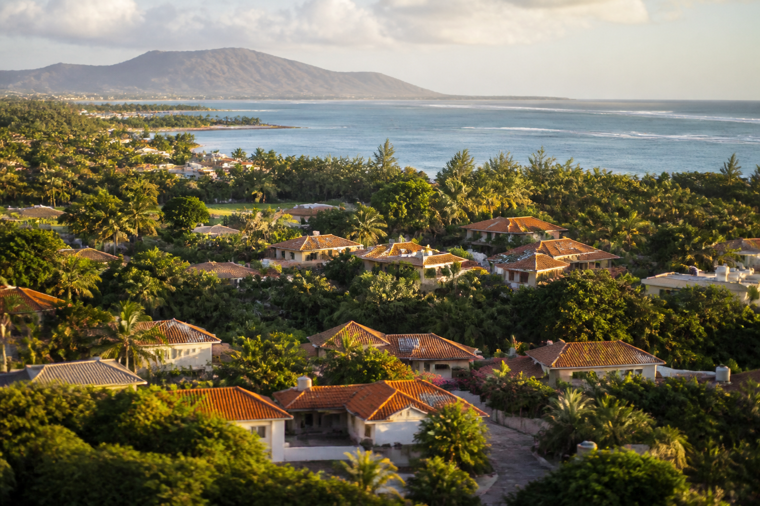 aerial view of a Mauritius coastal residential community near Tamarin with terracotta rooftops, dense tropical greenery, and turquoise lagoon visible in the background, golden afternoon light, no people